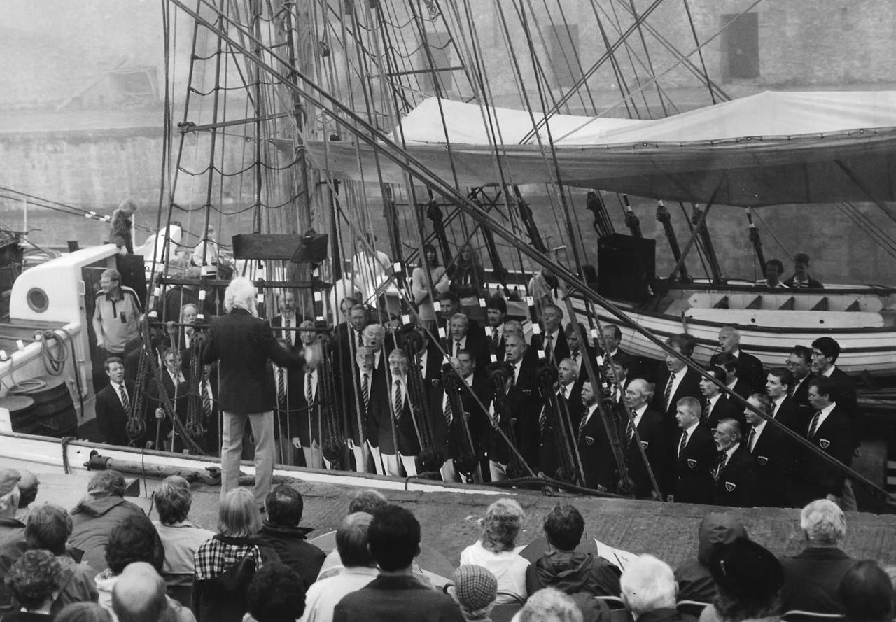 Black and white photo of the choir singing aboard a tall ship in harbour, watched by a crowd on the quay