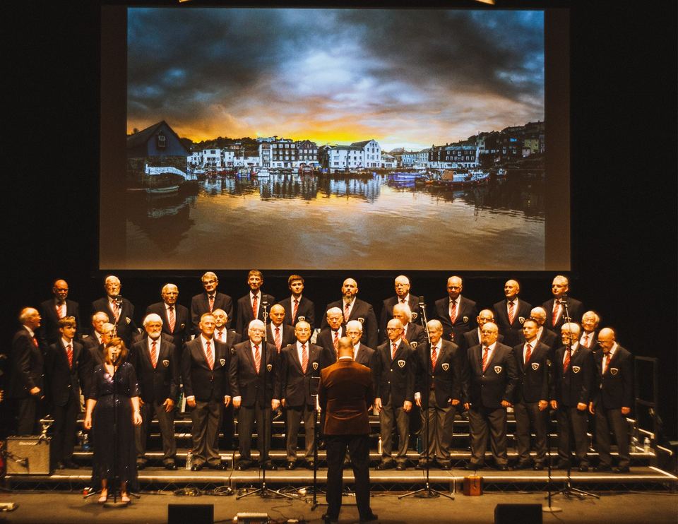 Mevagissey Male Choir on stage at Hall for Cornwall, with a photograph of Mevagissey harbour projected behind them