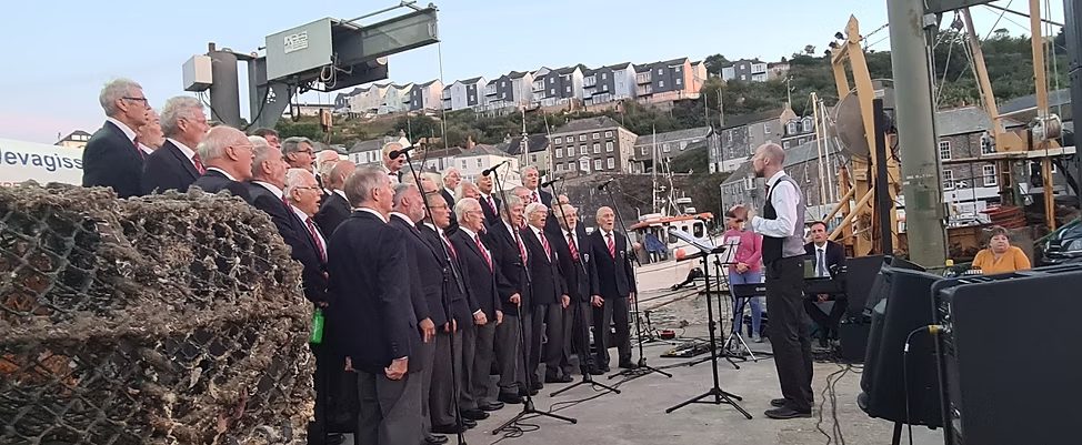 The choir singing on Mevagissey quay at dusk, with the harbour and the village behind them